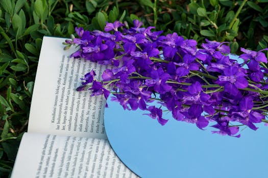 Purple flowers reflected in a circular mirror placed on an open book, set against green grass.